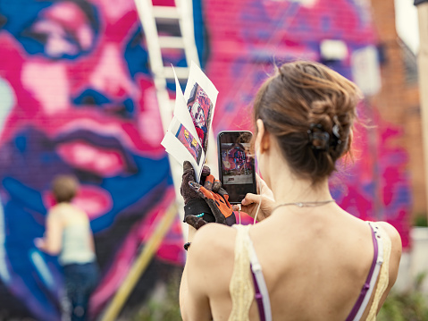 Young woman taking a photo with mobile phone of the mural she is creating with her friend in the background. She is dressed in casual work clothes. Exterior of urban street, back alley with old brick wall of the house.