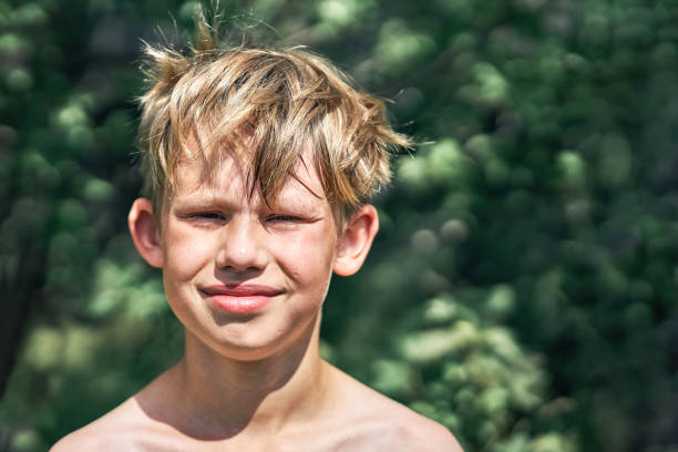 Smiling junior schoolboy with blond disheveled rough hair posing for camera in green park on nice summer day close view
