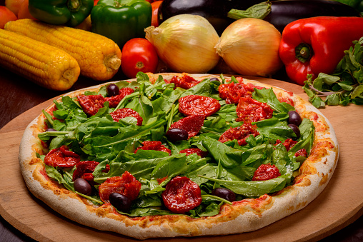 Arugula and sun dried tomato pizza on wooden board and vegetables in the background.