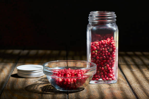 ciotola di vetro e barattolo di peperoni rosa su un tavolo di legno alla luce del sole, primo piano - pink peppercorn foto e immagini stock