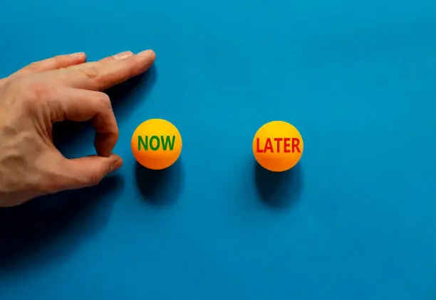 Photo of Now or later symbol. Male hand is about to flick the ball. Orange table tennis balls with words now later. Beautiful blue background. Business, now or later concept.