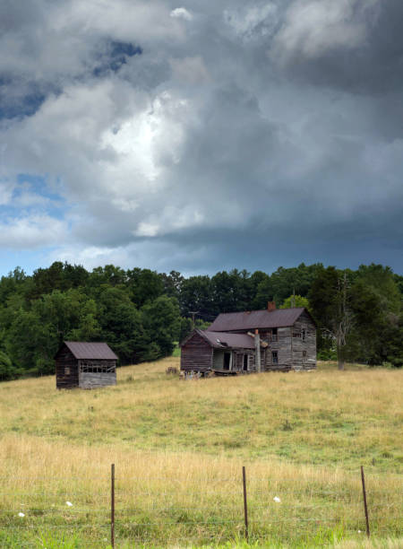 Dramatic skies overhead.  Old homestead in a field with wire fencing in the foreground.