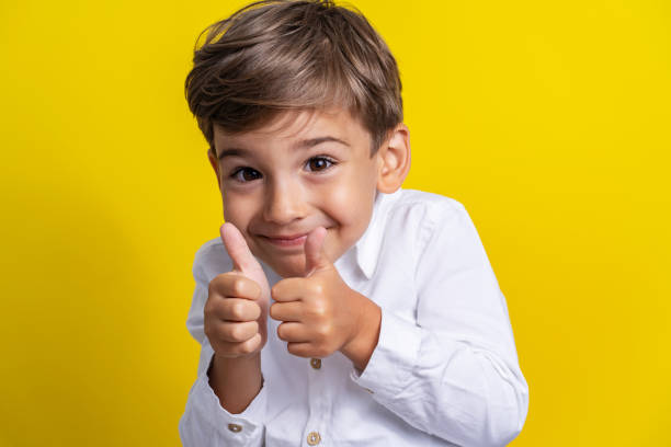 vista frontale del piccolo ragazzo caucasico di quattro anni in piedi di fronte allo studio di sfondo giallo girato sorridente e tenendo i pollici in su supporto per bambini e concetto di successo - 4 5 years foto e immagini stock