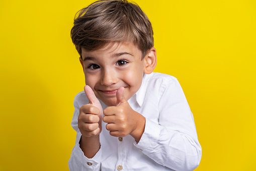 Front view of small caucasian boy four years old standing in front of yellow background studio shot smiling and holding thumbs up child support and success concept