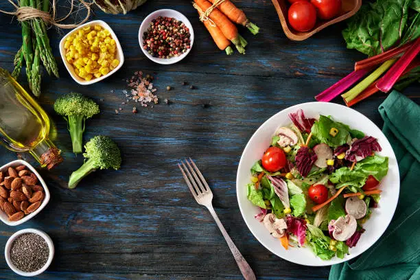 Top view of a colorful spring salad on rustic white wood table. Included ingredients: Olive oil, peppermint, chia, tomatoes, broccoli, lettuce, bell peppers, mushroom, carrots, radicchio and almonds.