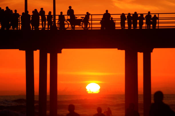 The sun sets in the background as beachgoers enjoy a beautiful sunset from the Huntington Beach Pier and the surrounding beaches.