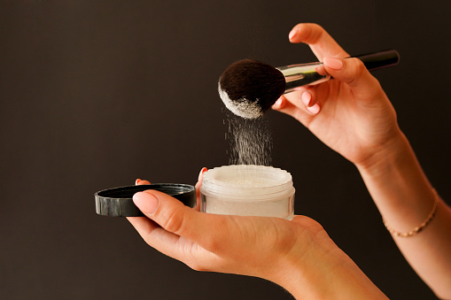 Female hands holding jar of powder and cosmetic brush on black background.