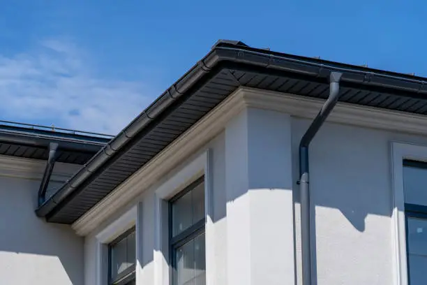 Corner of house with windows, new gray metal tile roof and rain gutter. Metallic Guttering System, Guttering and Drainage Pipe Exterior