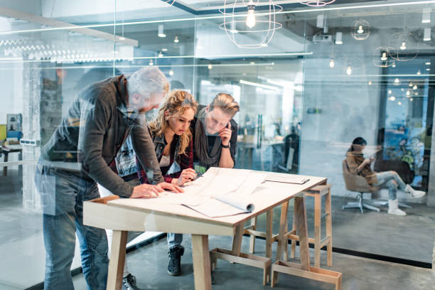 Building contractors examining blueprints in the office. Team of architects cooperating while analyzing blueprints on a meeting in the office. The view is through glass. architect office stock pictures, royalty-free photos & images