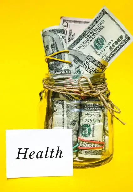 Photo of Glass jars filled with dollar bills, savings inside glass jar, money isolated on yellow background. Paper note written word HEALTH. Business budget of wealth