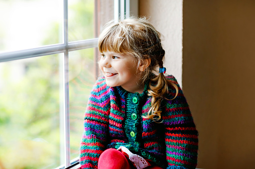 Little girl sitting by window. Preschool child wear cozy self knitted wool sweater. Toddler watching on rain outside. Autumn and winter season