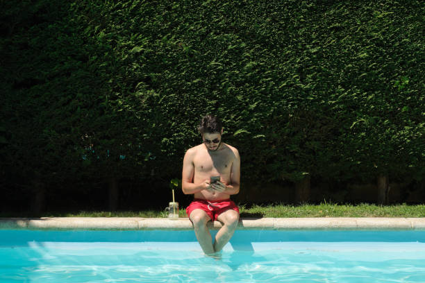 istock Young man wearing sunglasses using his smartphone sitting on the swimming pool edge. Summer concept. 1328455730