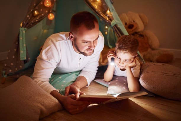 istock Reading book and using flashlight. Father and his young young spending time together indoors near tent 1328458244