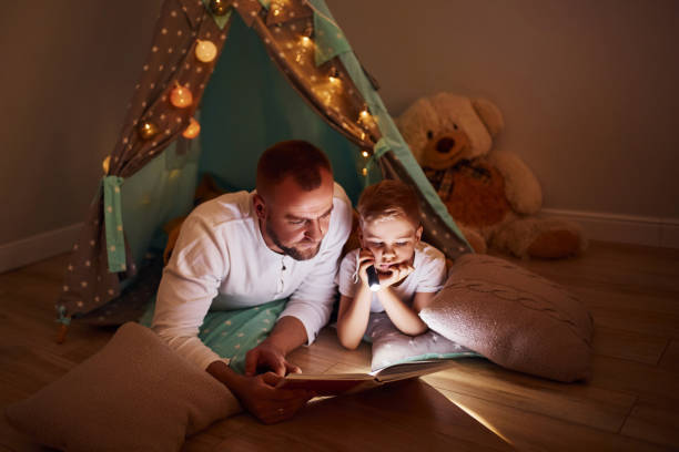 istock Reading book and using flashlight. Father and his young young spending time together indoors near tent 1328458271