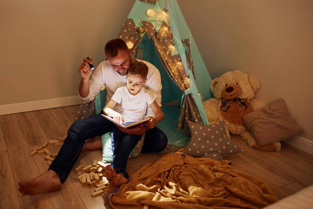 istock Reading book and using flashlight. Father and his young young spending time together indoors near tent 1328458310