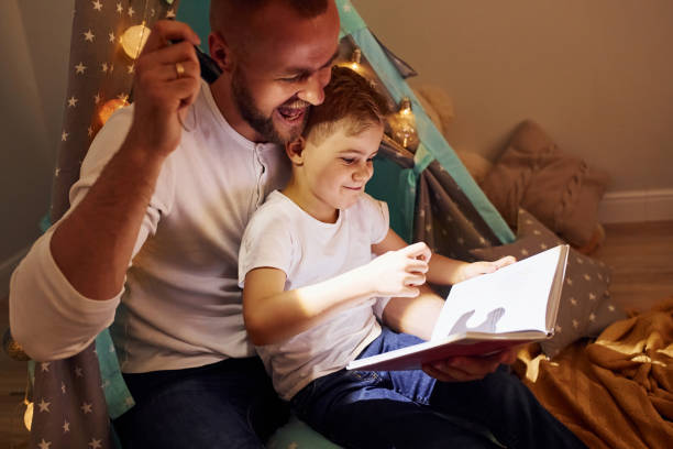 istock Reading book and using flashlight. Father and his young young spending time together indoors near tent 1328458311