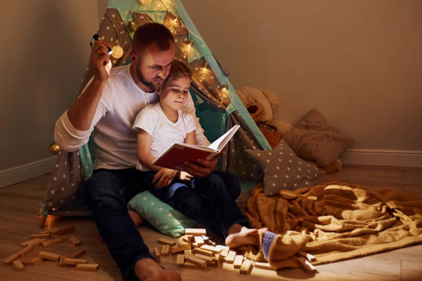 istock Reading book and using flashlight. Father and his young young spending time together indoors near tent 1328458336