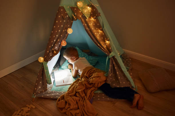 istock Reading book and using flashlight. Young boy in casual clothes lying down near tent at evening time 1328458498