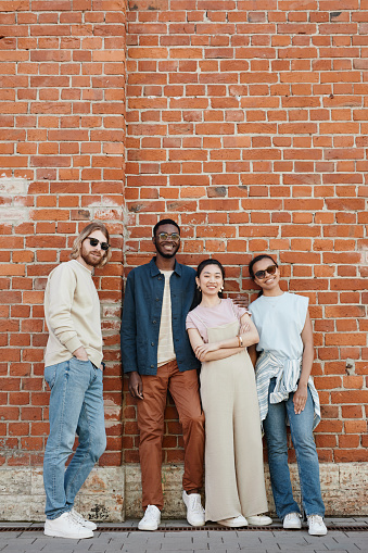 Full length view at diverse group of young people posing outdoors and looking at camera while standing by brick wall in urban setting, copy space