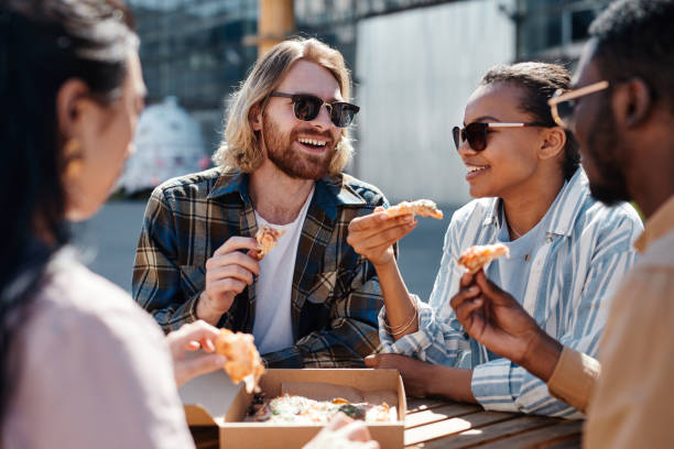 Group of Friends Eating Pizza Outdoors Diverse group of carefree young people enjoying pizza outdoors, scene lit by sunlight, copy space black people eating stock pictures, royalty-free photos & images