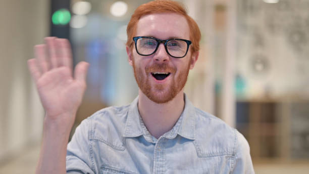 istock Cheerful Young Redhead Man Waving at the Camera 1328470929