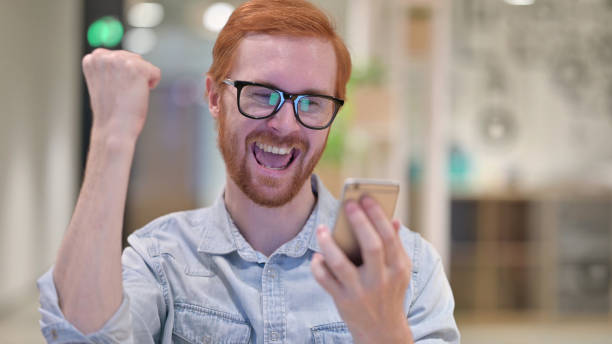 istock Young Redhead Man Celebrating Success on Smartphone 1328471657