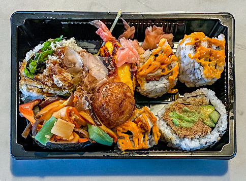 Close-up directly above: selection of sushi pieces for lunch in black plastic take-away tray with wasabi: maki, California roll,