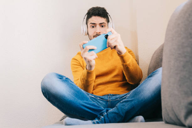 自宅のソファに座って、興奮した表情で携帯電話でバーチャルリアリティゲームをプレイするヘッドフォンを持つ30歳の男性。 - portable player ストックフォトと画像