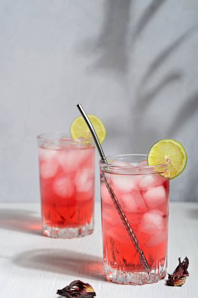 Two glasses of cold red hibiscus tea served with metal straw, ice cubes and lime slice under summer sunlight with plant shadows Cold refreshing hibiscus herbal tea made from magenta pink colored flowers served in drinking glass with ice cubes, lime slice and metal straw on white wooden table with summer sunlight and shadows pink tea stock pictures, royalty-free photos & images