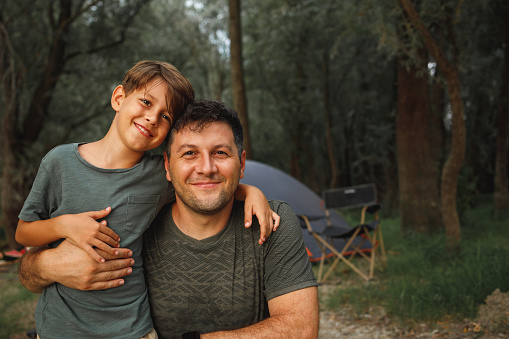 A smiling father and son hug and pose for the photo in front of a tent while camping in nature.