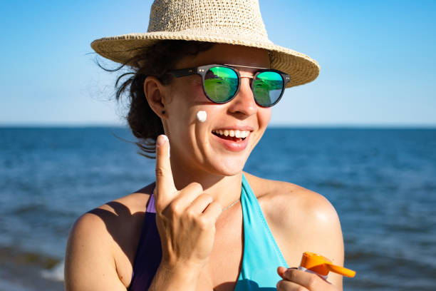 istock Suntan spf lotion. Beautiful woman applying sunscreen solar cream from a plastic container to her cheek, wearing straw summer hat and blue swimming suit with sea in background. Sunscreen protection. 1328491426