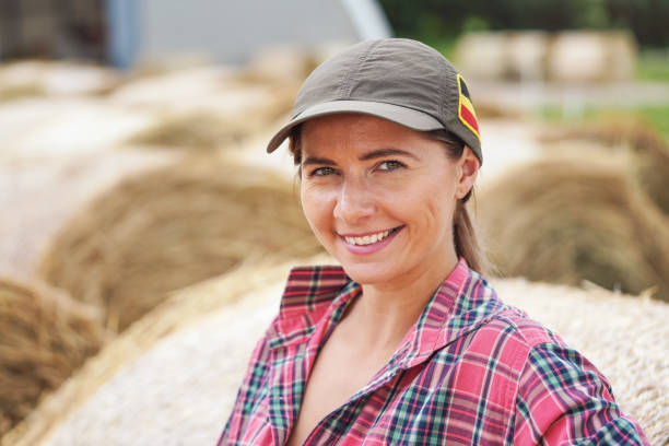 istock Young woman in shirt and cap smiling, hay stack rolls behind her 1328491544