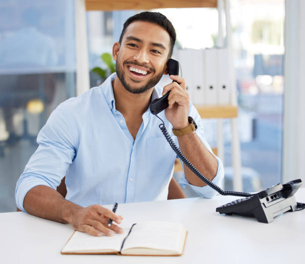 foto de um jovem empresário escrevendo em um caderno enquanto usava o telefone no trabalho - telefone fixo - fotografias e filmes do acervo