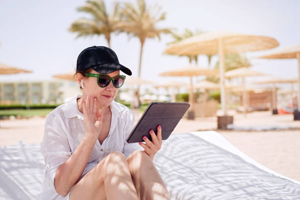 istock Mature woman having a video chat through tablet on background the beach. Remote work, summer vacation concept, beach work, freelance work and leisure 1328496198