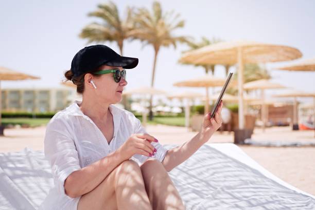 istock Mature woman having a video chat through tablet on background the beach. Remote work, summer vacation concept, beach work, freelance work and leisure 1328496199