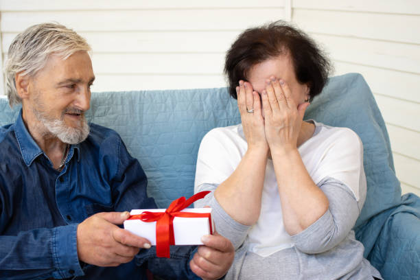 homem de cabelos grisalhos sênior segurando caixa de presente nas mãos e fazendo surpresa para a esposa, enquanto mulher madura cobrindo seus olhos. parabéns e comemoração de aniversário ou aniversário, conceito de dia dos namorados - men latin american and hispanic ethnicity southern european descent mature adult - fotografias e filmes do acervo