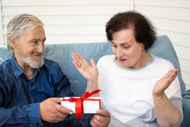 surprise. excited senior woman receiving gift box with red bow from her loving mature grey haired husband, sitting on sofa at home terrace.celebration of birthday or anniversary,valentine day concept - men latin american and hispanic ethnicity southern european descent mature adult imagens e fotografias de stock