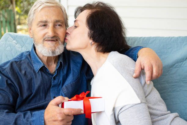 idosa beijando seu marido maduro de cabelos grisalhos, obrigado pelo presente surpresa recebido dele, sentado no sofá do terraço ao ar livre. feliz aniversário ou aniversário, conceito do dia dos namorados - men latin american and hispanic ethnicity southern european descent mature adult - fotografias e filmes do acervo