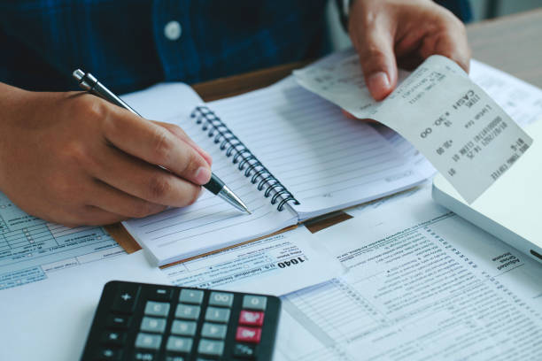 homme utilisant calculer les factures domestiques sur un bureau en bois dans le bureau et les affaires fond de travail. jeune homme vérifiant l’équilibre et les coûts avec le formulaire irs 1040 des états-unis, la fiscalité, les statistiques et les  - to accumulate photos et images de collection