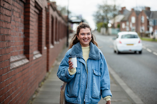 A medium close-up of a young woman wearing a denim jacket walking towards Work. She is carrying a disposable coffee cups and is smiling as she walks towards the town centre she's also carrying a bag over his shoulder as she walks down the road.