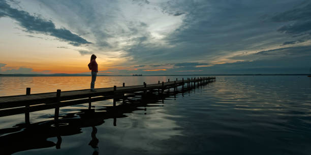 silhouette di donna matura in piedi sul molo al lago con maestoso paesaggio nuvoloso al crepuscolo - steinhuder meer foto e immagini stock