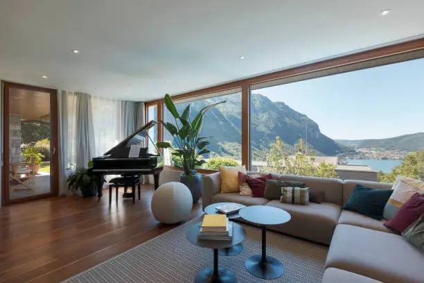 Living room with large light-colored sofa and lots of cushions and a black piano. Large window overlooking the valley with a lake view. Nobody inside