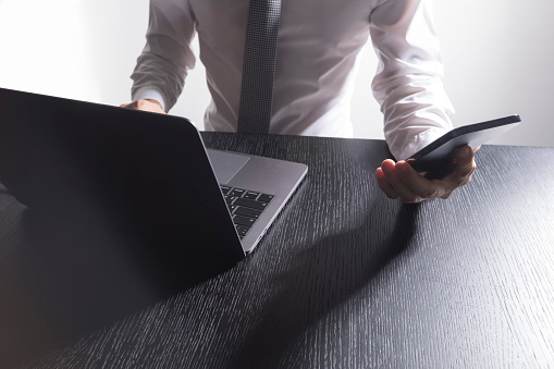 Close-up of the hands of a well-dressed man in a white shirt looking at his smartphone, using a laptop on a desk.