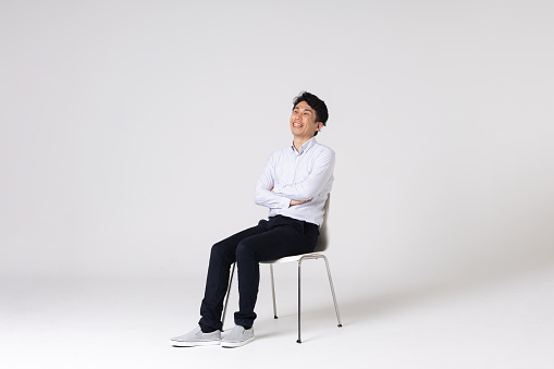 Full-length portrait of an Asian man on a white background.
Full body shot. Japanese male, 30s.
Healthy and confident figure.
sitting on a chair. holding a smart phone. smiling.
Looking at camera.
Shot in a studio with a white background.
Horizontal shot.
