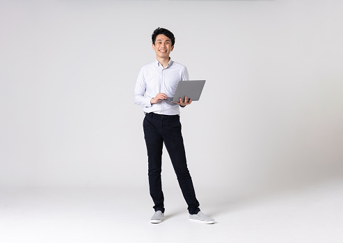 Full-length portrait of an Asian man on a white background.
Full body shot. Japanese male, 30s.
Healthy and confident figure.
smiling. Looking at camera. Standing.
Holding a laptop.
Shot in a studio with a white background.
Horizontal shot.