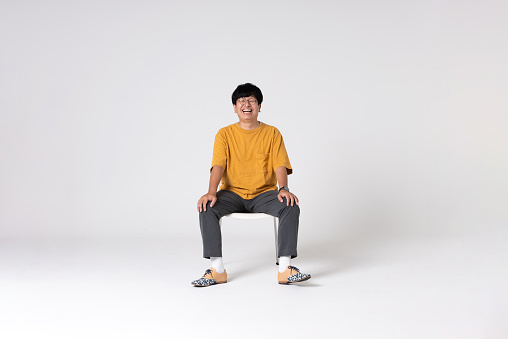 Full-length portrait of an Asian man on a white background.\nFull body shot. Japanese male, 30s.\nHealthy and confident figure.\nsitting on a chair. smiling.\nLooking at camera.\nShot in a studio with a white background.\nHorizontal shot.