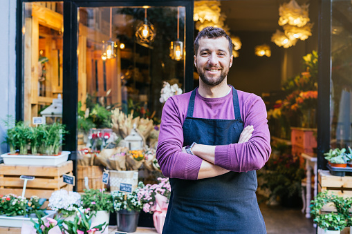 El dueño de una floristería en un delantal tiene los brazos cruzados frente a su tienda de flores. Tiene un delantal oscuro, está sonriendo. photo