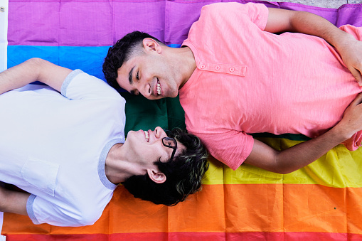 two Latino gay men lying on a rainbow flag, looking at each other smiling