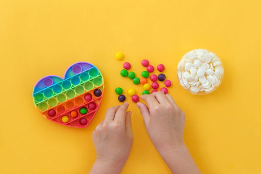 Use the popular pop it toy as a chocolate mold. Children is hands put colorful candies in a popit heart-shaped silicone mold on a yellow background. DIY concept. Step-by-step photo instructions. Step 2.