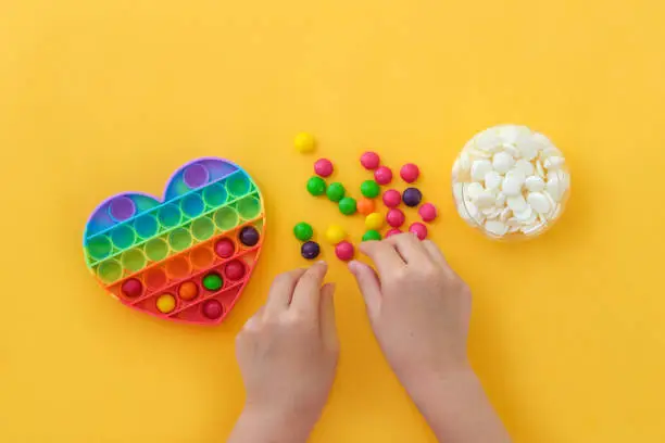 Photo of Use the popular pop it toy as a chocolate mold. Children is hands put colorful candies in a popit heart-shaped silicone mold on a yellow background. DIY concept. Step-by-step photo instructions. Step 2.